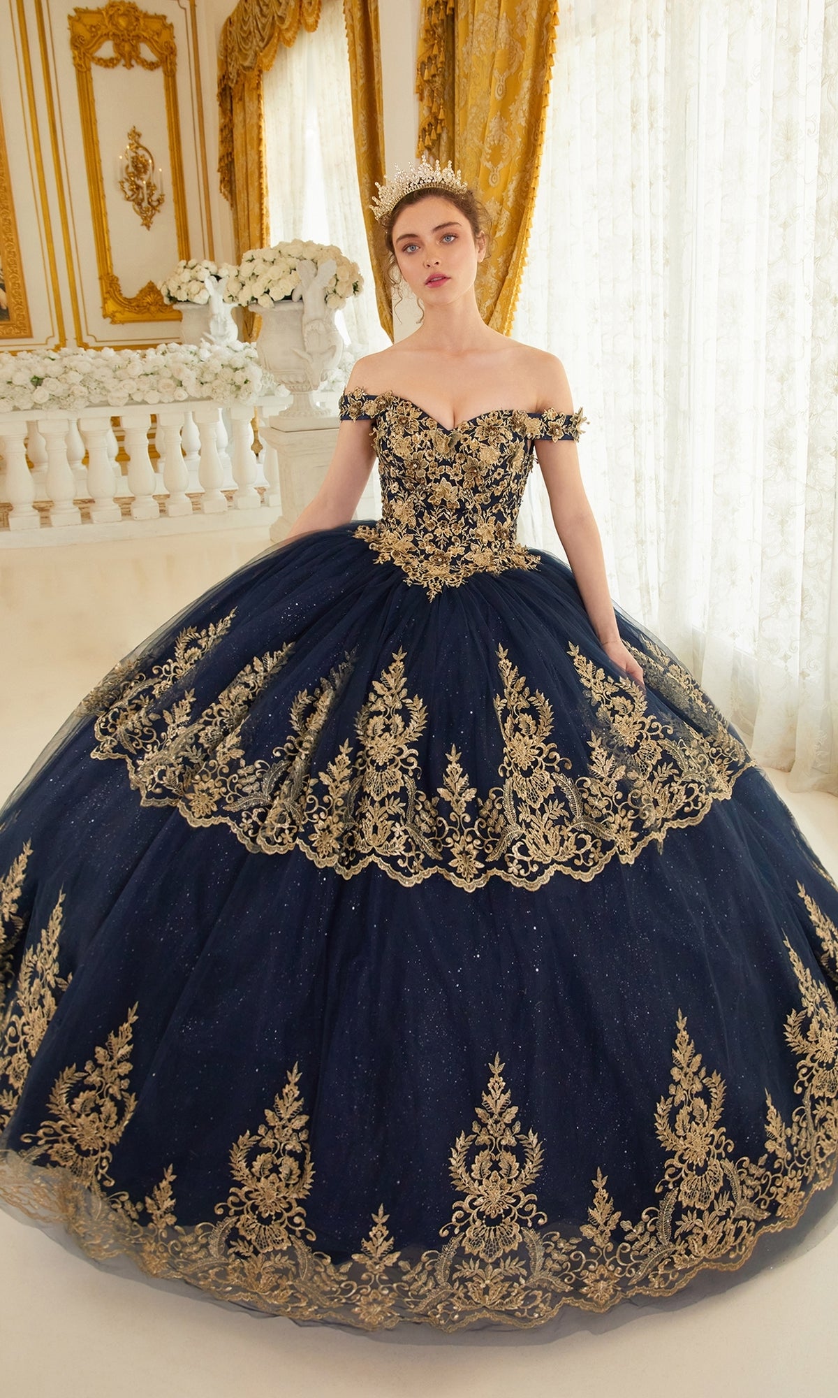 A woman in the Ladivine 15705 Quince Dress with gold embroidery and a tiara stands in an ornate, gold-accented room adorned with white flowers and flowing curtains.
